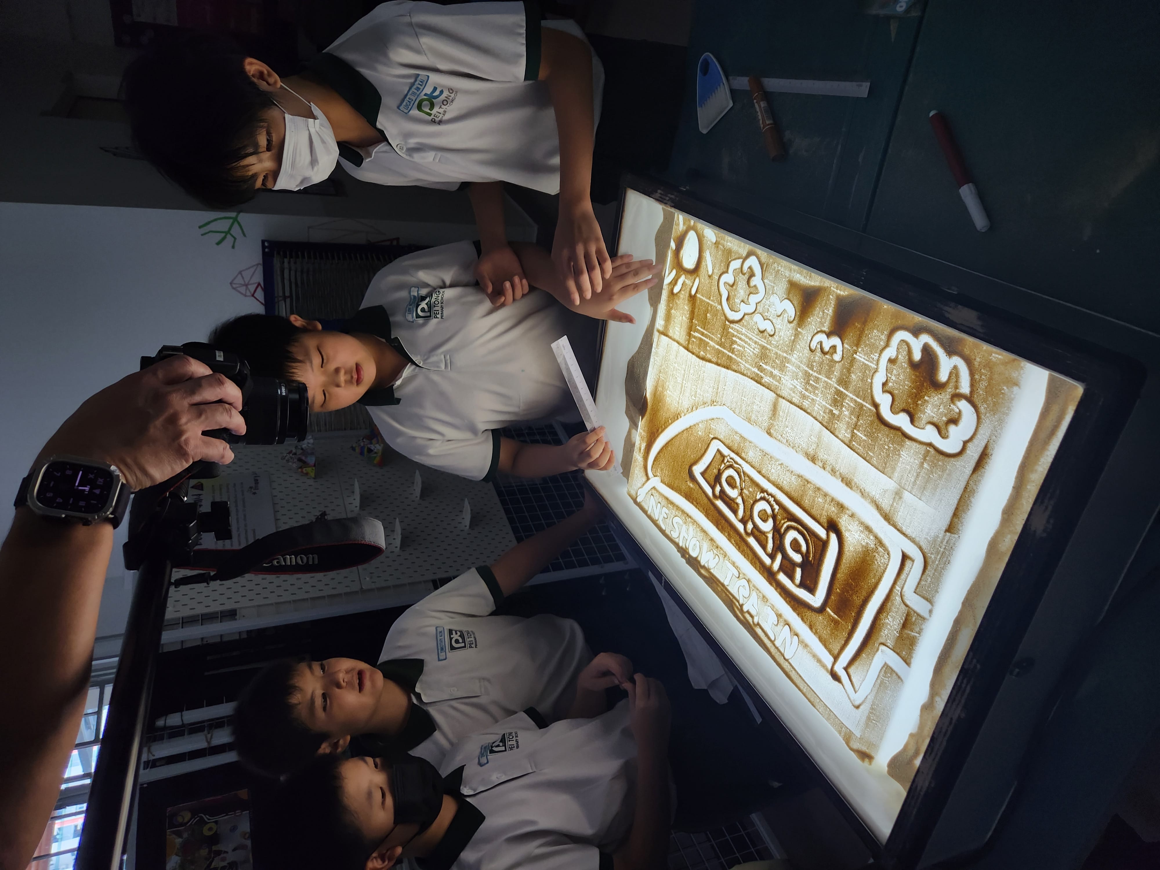 Students gather around a light table, creating and observing a sand artwork while an instructor photographs their work during a sand animation art lesson.